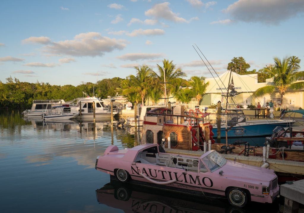 A bubblegum pink convertible in the water, like a boat, at a bar on the edge of the water.