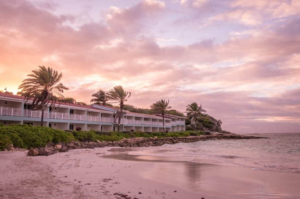 A pink sunset over a beach with white hotel rooms and palm trees in the background.