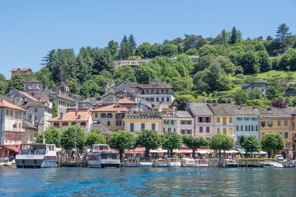 Pastel-colored houses are sitting right on Lake Orta, with boats in front of them in the water. A large green hill rises behind them underneath a bright blue sky.