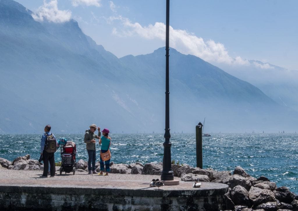 Four adults and a baby carriage stand on a jetty jutting out into the blue Lake Garda, where a windy day is stirring up white caps in the waves. The mountains are jagged in the background and seem to be falling into the lake.
