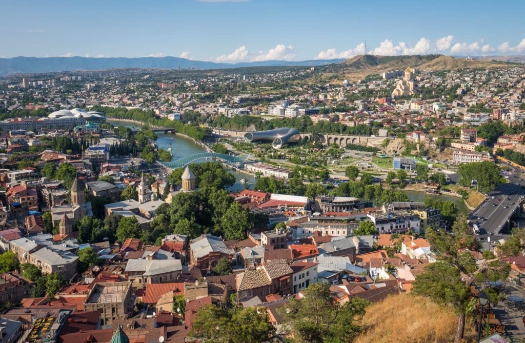 A gorgeous view of Tbilisi underneath a blue sky, buildings with orange roofs, the green river snaking through the city, a few of the modern glassy buildings poking up in the photo.