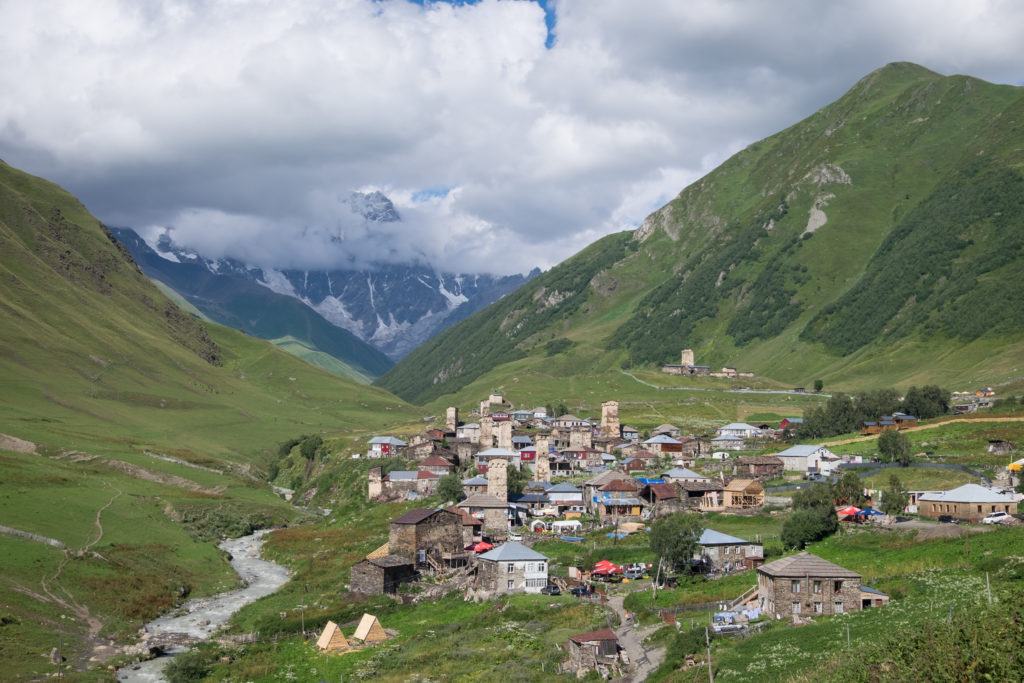 A distance view of Ushguli, with the stone towers of the village towering over the green landscape but not the surrounding green mountains.