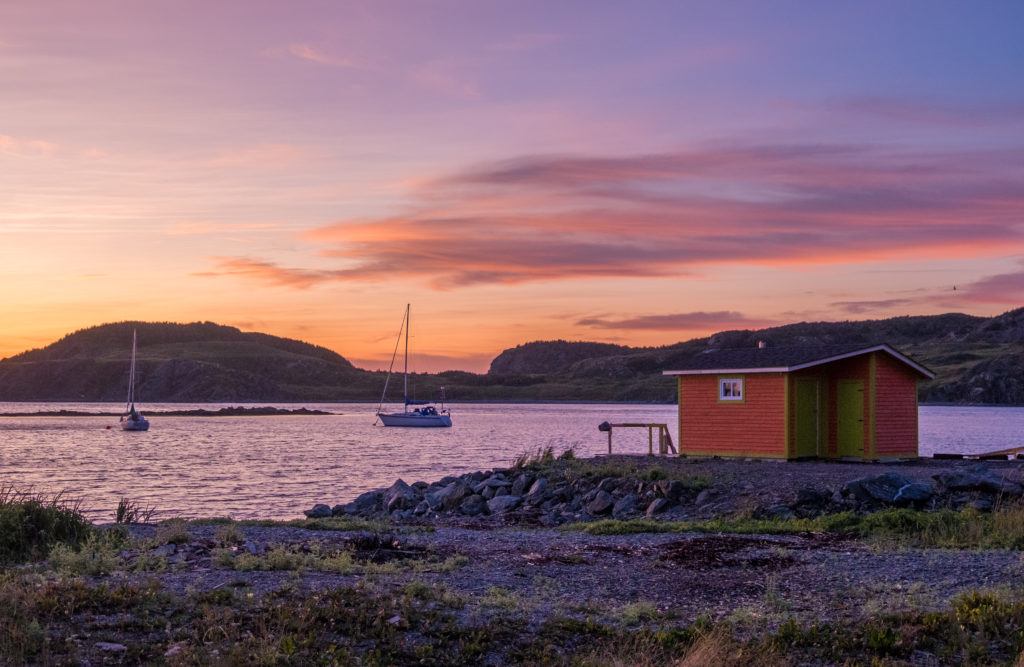 A sunset with a light purple sky and pink and dark purple clouds, in front of a red fishing hut and a sailboat