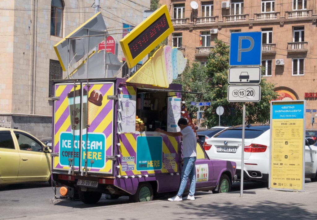 A man ordering from a yellow and purple-striped food truck on a street in Yerevan.