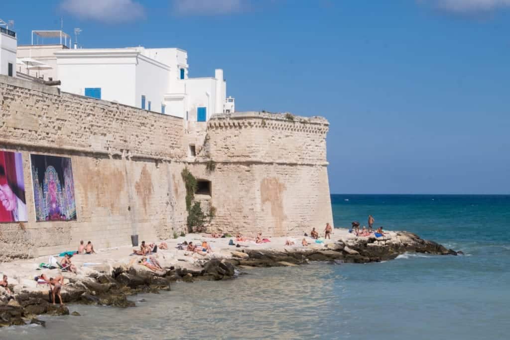 A city wall juts into the ocean; people sun themselves on the rocks surrounding the wall.