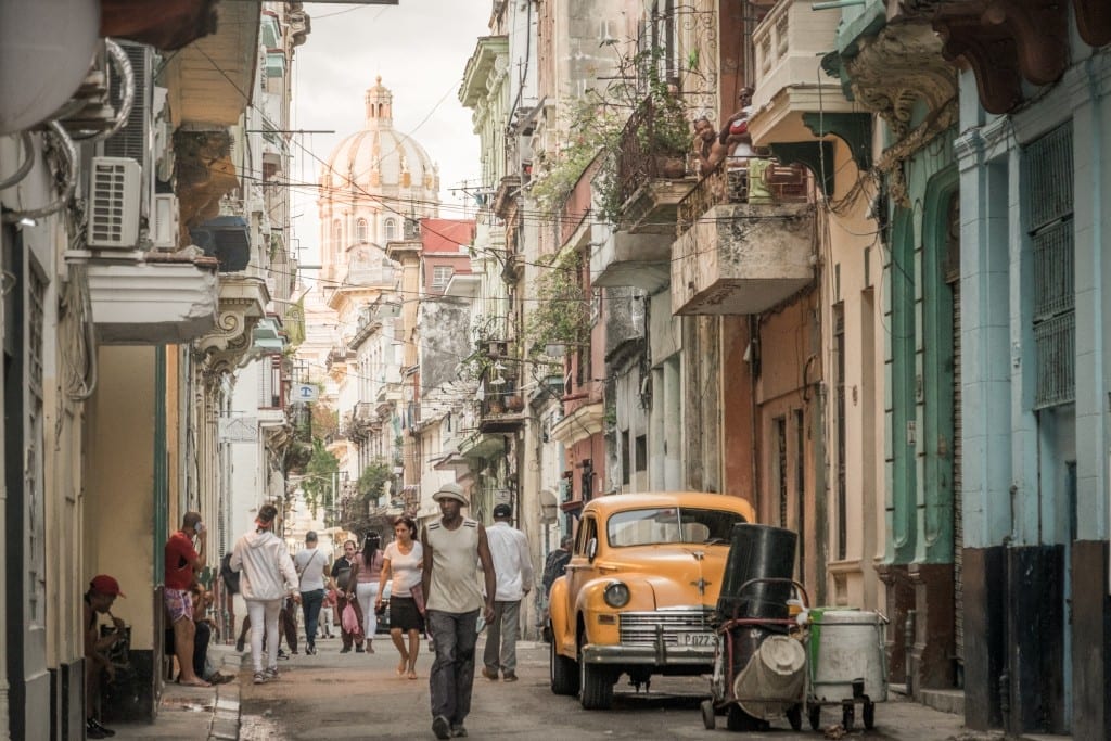 Chaotic Havana streets filled with people, a yellow car, and a church tower in the background.