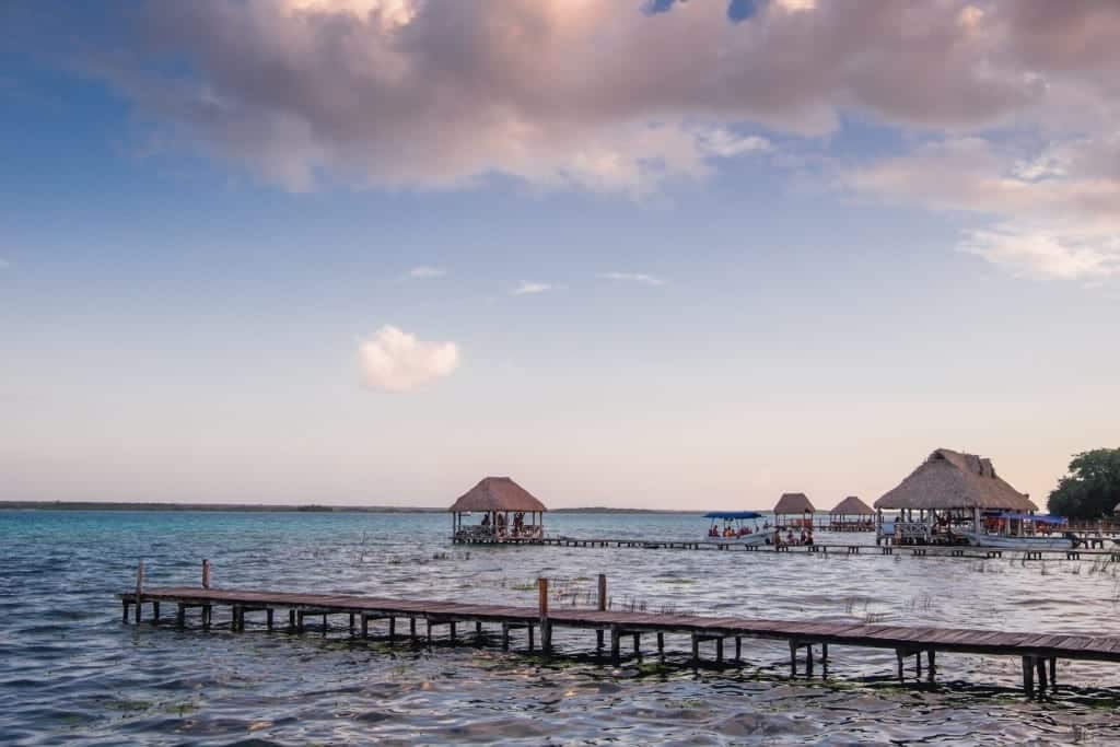 Huts on Bacalar Lagoon underneath a pink and blue sky.