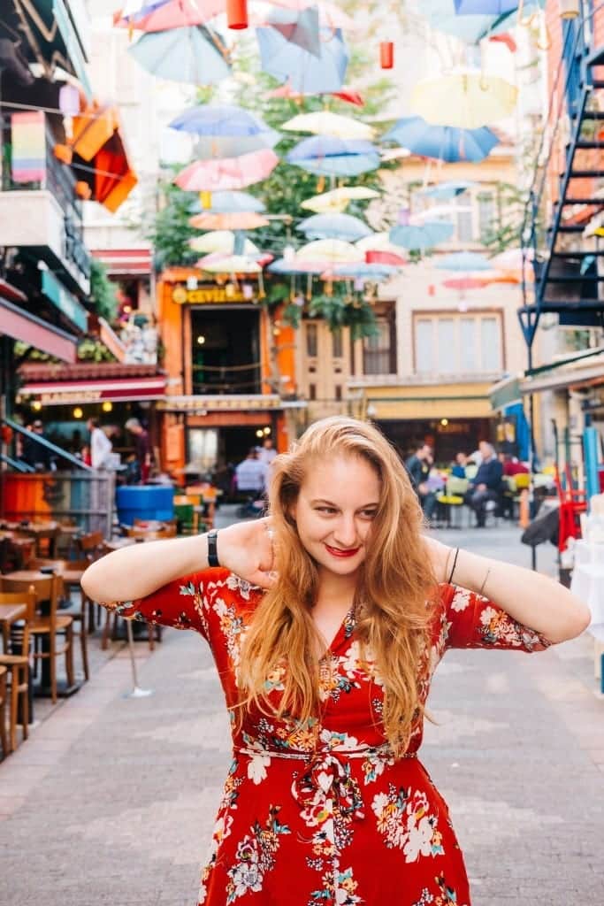 Katie wearing a red dress and smiling on an Istanbul street with bright shops and hanging umbrellas.