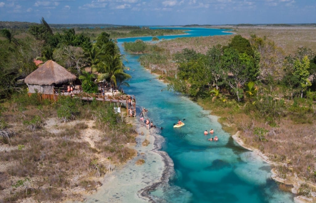 An aerial view of Los Rapidos: a turquoise river going through the green landscape, a small hut on shore, people swimming in the river.