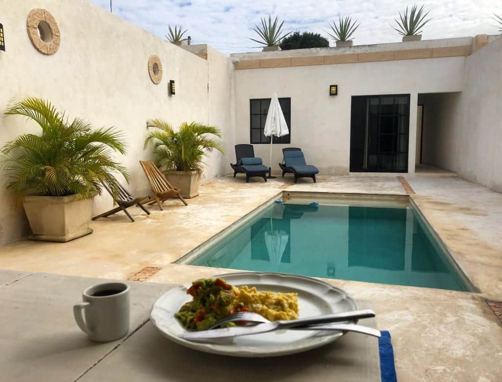 Our courtyard at our house in Merida, Mexico, with a cement floor, a turquoise pool, and large planters with green ferns in them. In the foreground is a plate of scrambled eggs and a coffee on the table.