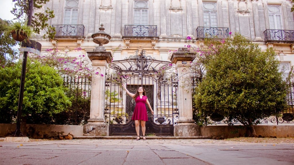 Kate wears a red dress and stands in front of a wrought-iron gate to a mansion, surrounded by purple flowers, in Merida, Mexico
