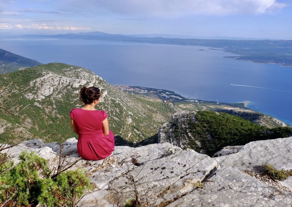 Kate wears a berry-colored dress and sits along on top of a cliff, looking over green mountains and the ocean, Hvar Island in the distance, in Bra?.