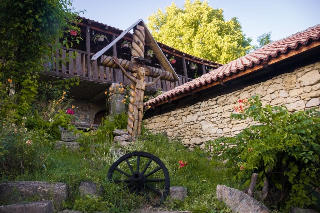 A hand-carved wooden crucifix in the middle of a collection of stone and wood buildings and flowers in the Serbian countryside.