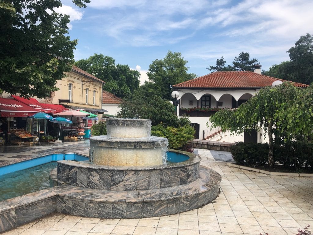 A layered cake-shaped fountain in the square of Sokobanja, shops and white buildings in the distance.