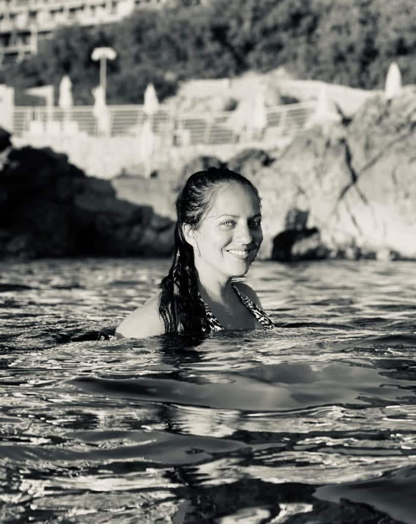 Kate swims in the ocean in Dubrovnik, Croatia, the water reflecting her image in the waves, rocky cliffs behind her.