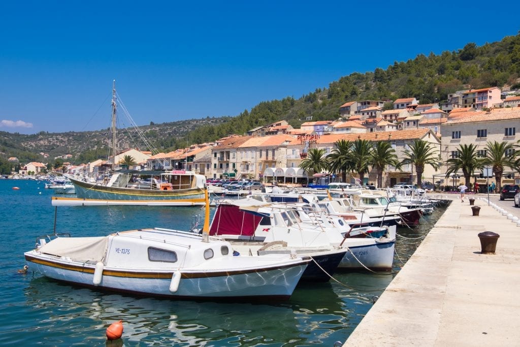 Vela Luka: a row of boats anchored to the waterfront, palm trees and orange-roofed houses in the background.