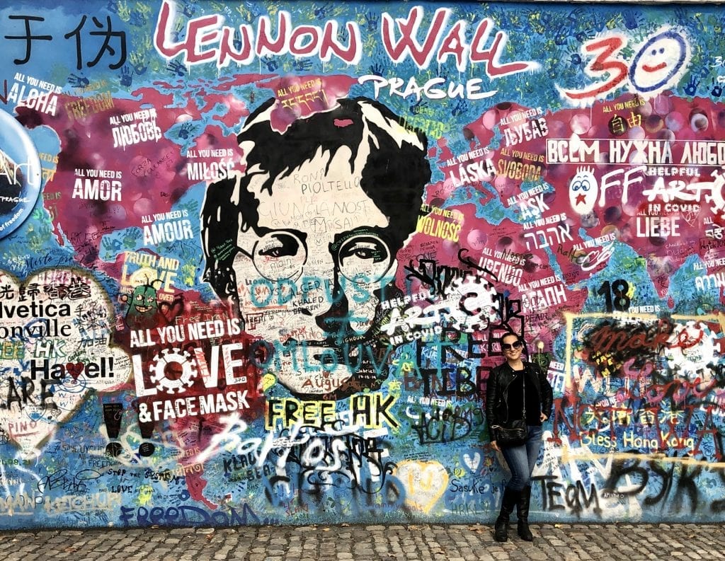 Kate stands in front of the Lennon Wall in Prague, featuring an image of John Lennon and lots of graffiti on top of a map. Part of it says "All you need is Love and Face Mask." Kate wears a leather jacket, skinny jeans, tall black boots, big black sunglasses.