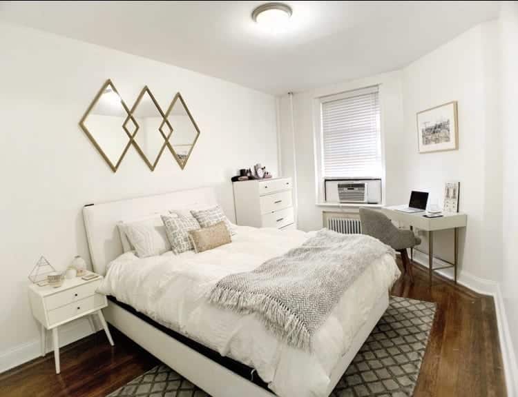 Kate's New York bedroom: a large white bed with a white bedspread, silver and gold pillows, a gray and white blanket, white nightstand, dresser, and small desk, gray rug, gray and white striped office chair, and gold mirror shaped with three diamonds hanging above the bed.