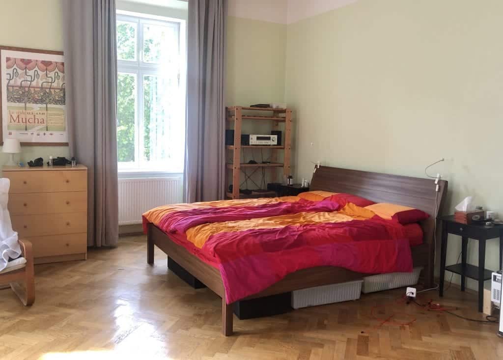 A wooden bed covered with a blocky red pink and orange IKEA bedspread, open shelving and dresser in the background.