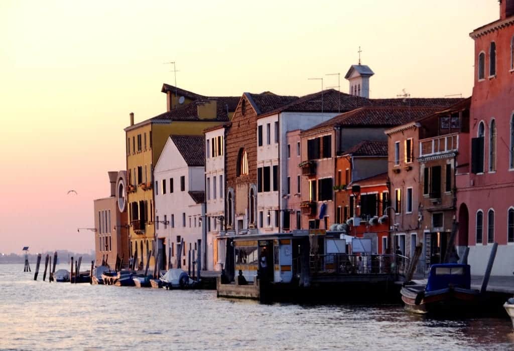 View from a canal of Venetian homes built along the water, turning a pink shade at sunset.