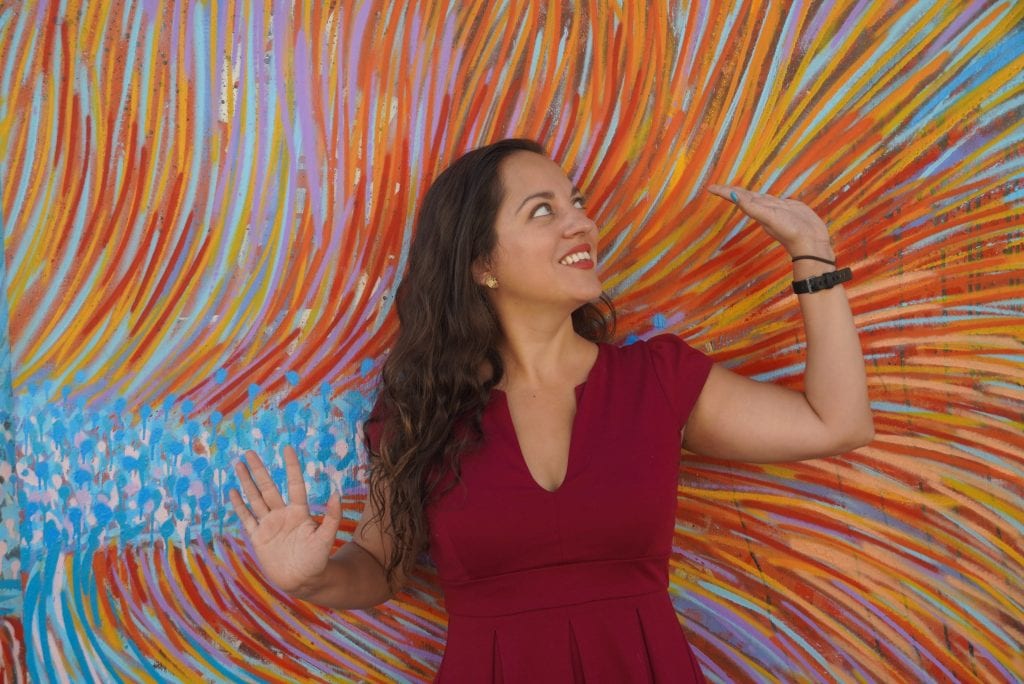 Kate posing with her arms in the air in front of a mural of many different colors in an abstract design.