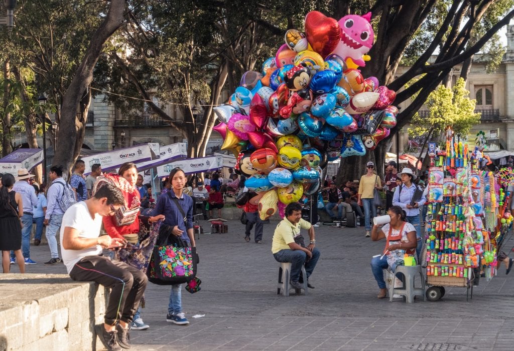 People walking down the main square of Oaxaca, the zocalo. One man carries a huge bundle of balloons; other people are sitting and looking at their phones.