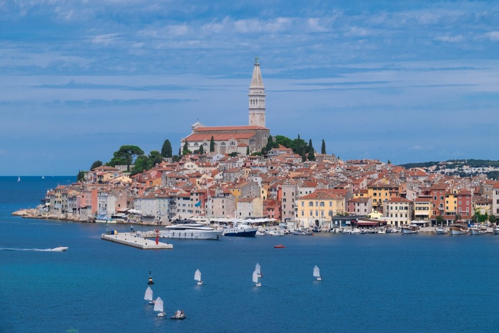 The Old Town of Rovinj, yellow and red-colored shuttered buildings with terra cotta roofs, a church steeple sticking straight up in the center, underneath a blue sky. Sailboats out in front of the old town.