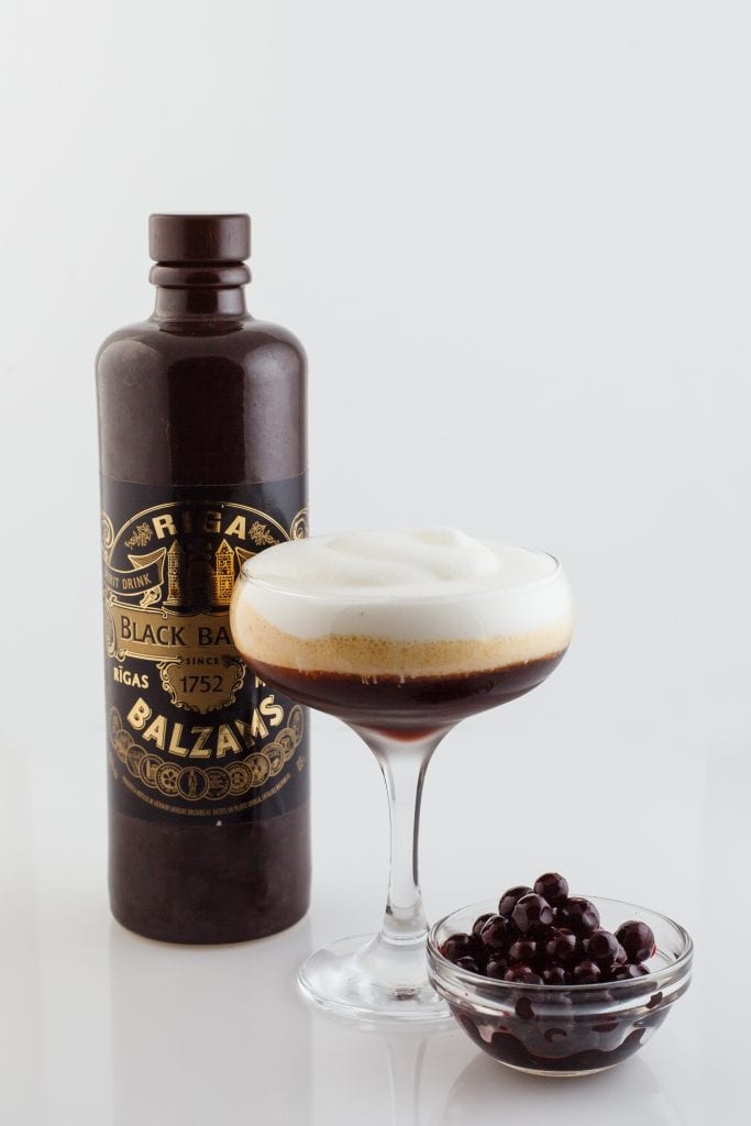 A dark brown bottle of Black Balsam liqueur with a black and gold label, a cocktail with a frothy white top, and a small bowl of dark brown berries.