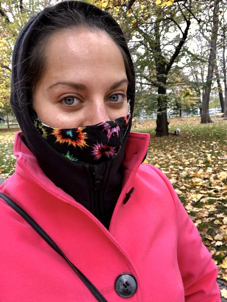 Kate selfie wearing a bright pink peacoat, black hooded sweatshirt and black and multicolor face mask, trying to stay warm on a cold day.