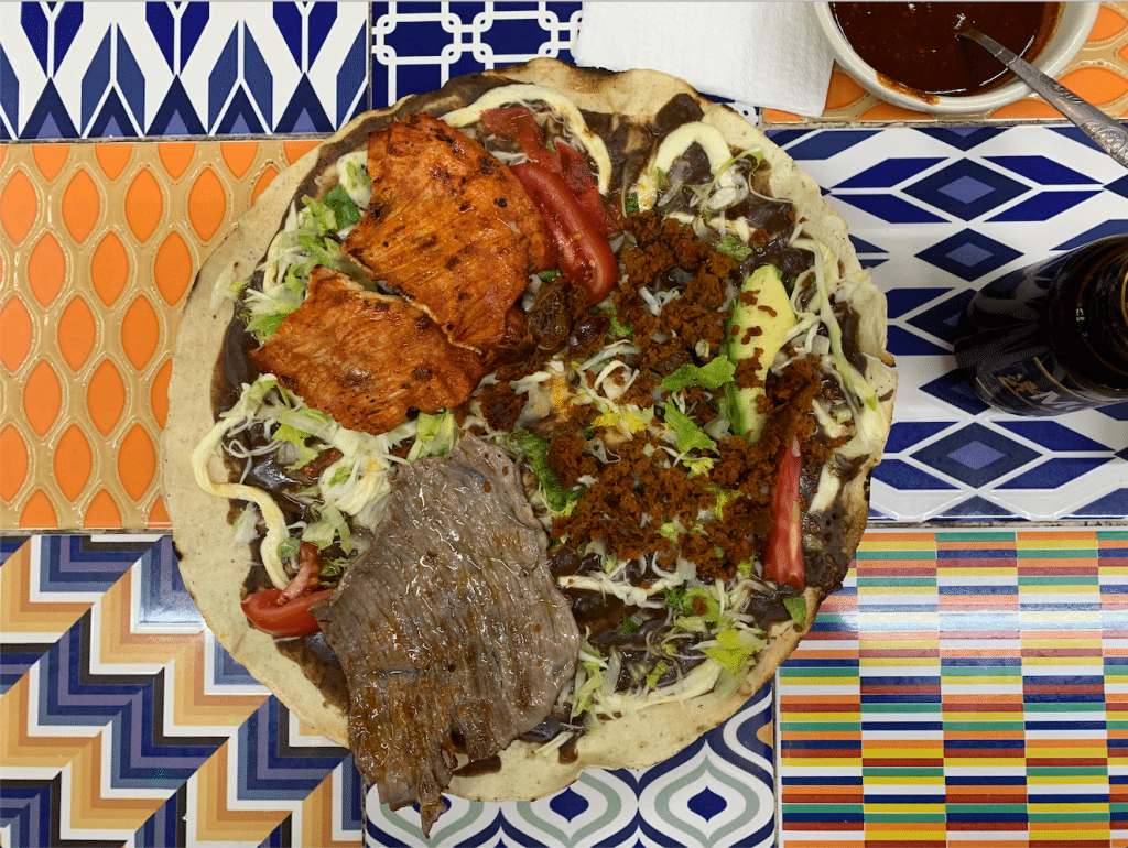 A Oaxacan tlauyda -- a giant tortilla topped with meat and vegetables, served on a blue and orange patterned tile table.