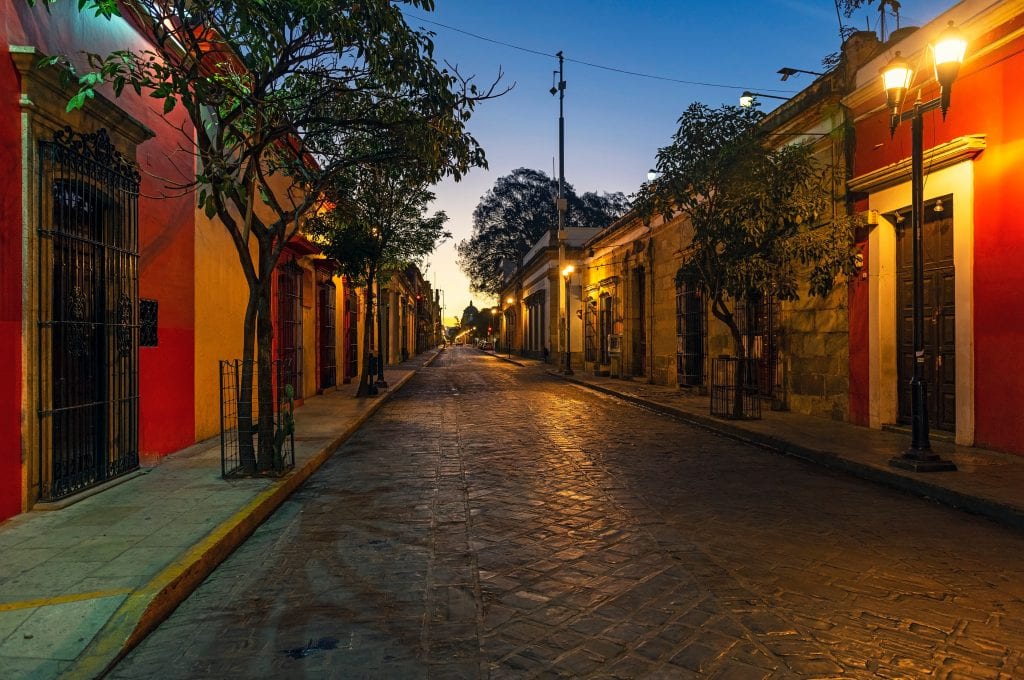 Oaxaca at dawn: right red buildings edged with trees, cobblestone street, lamps illuminating the colors, the sky blue and turning yellow.
